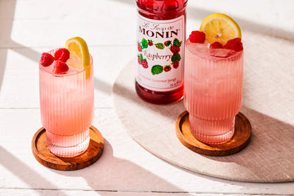 Monin Raspberry Syrup bottle beside refreshing raspberry lemonade garnished with fresh raspberries and lemon