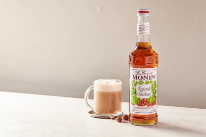 Monin Roasted Hazelnut Syrup bottle beside a creamy hazelnut latte