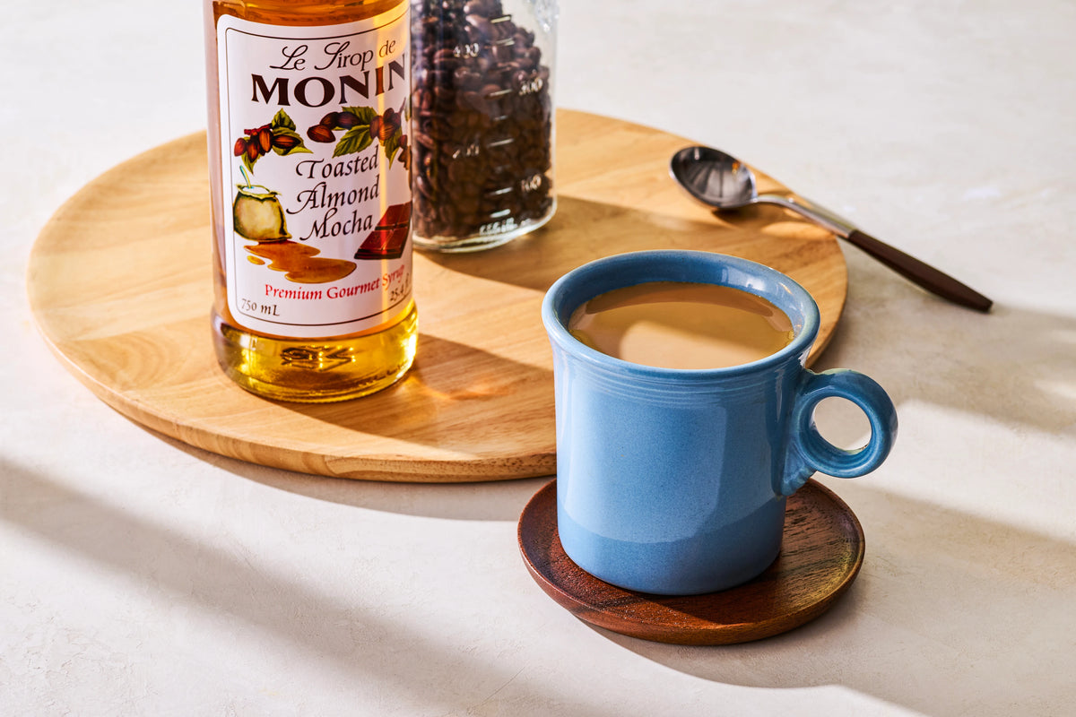 Monin Toasted Almond Mocha Syrup with a hot coffee in a blue mug and coffee beans