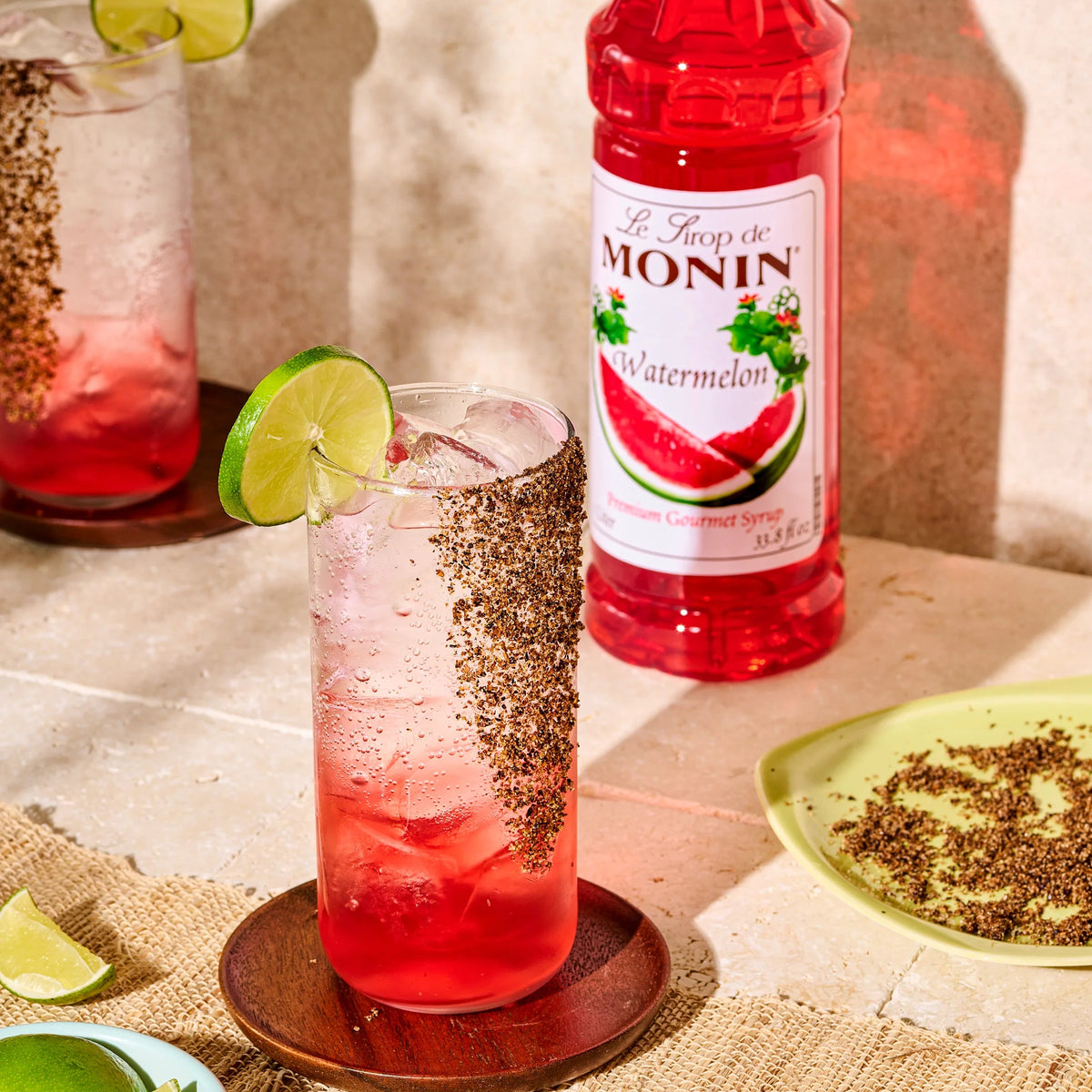 Monin Watermelon Syrup beside two sparkling watermelon drinks garnished with lime and a spiced rim