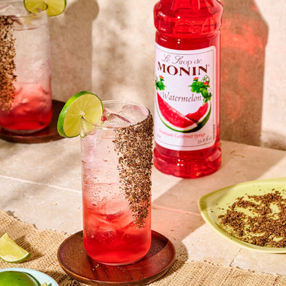 Monin Watermelon Syrup beside two sparkling watermelon drinks garnished with lime and a spiced rim